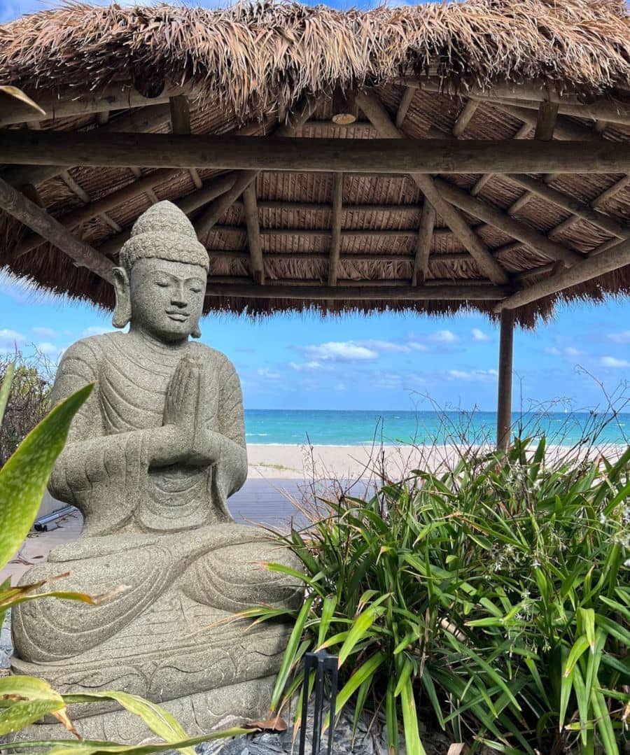 Concrete statue in front of a tiki on a beach, looking out to sea.